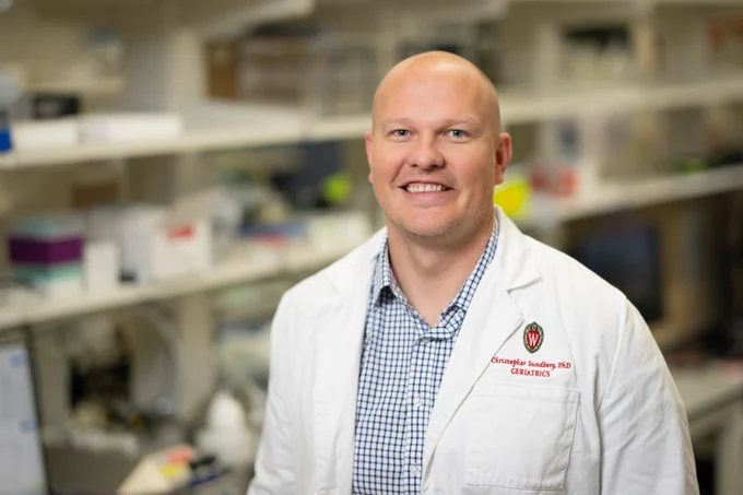 Dr. Christopher Sundberg wearing a white lab coat with a University of Wisconsin logo standing in a busy laboratory setting.