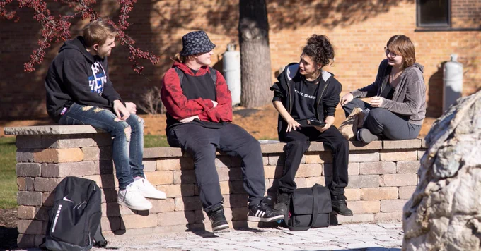 Four individuals are sitting on a low brick wall outdoors, engaged in a conversation. They are dressed in casual attire, with backpacks placed nearby, suggesting a school or college setting.