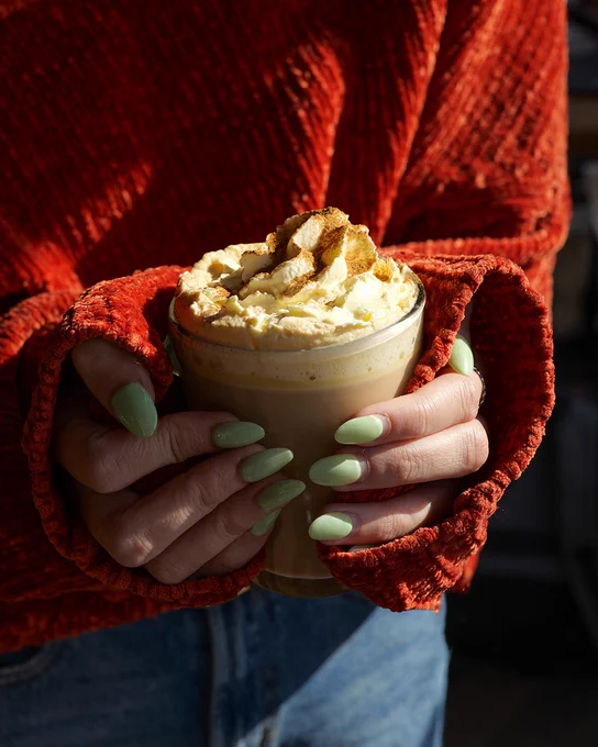 A person holding a glass of pumpkin spice latte topped with whipped cream and a sprinkle of spices. The person is wearing a textured orange sweater and has manicured nails with pale green polish.
