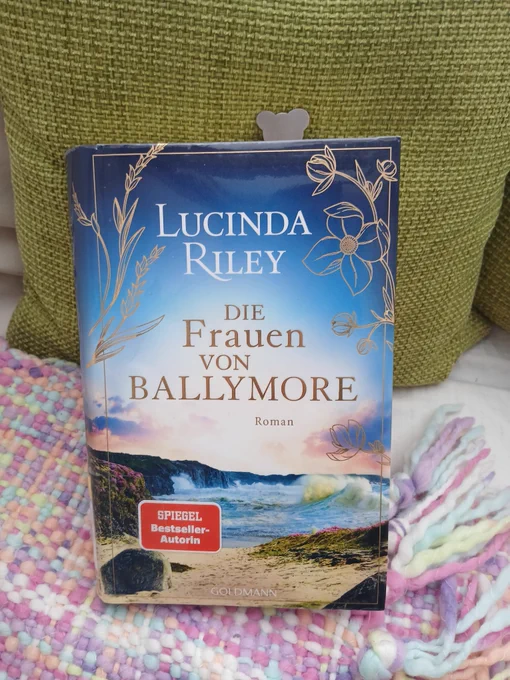 gebundenes Buch auf bunt gemusterter Wolldecke und moosgrünem Kissen. Oben guckt ein Lesezeichen aus Metall  in Form eines Bärenkopfes raus.
Lucinda Riley, Die Frauen von Ballymore

