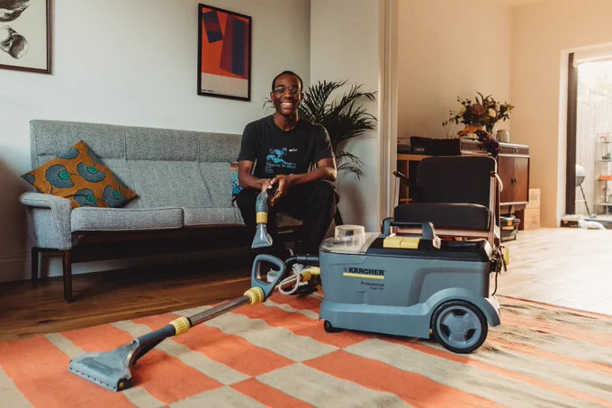 A man sitting on his sofa in his living room, posing happily for a photo. In front of him is the Library of Things' Karcher carpet cleaner