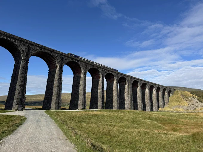 A long stone railway viaduct with multiple tall arches stretches across a grassy rural landscape under a bright blue sky, with a train traveling along the top of the bridge.