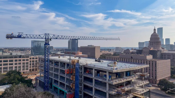 Aerial view of Phase II construction at the Texas Facilities Commission Capitol Complex in Austin, Texas, showing a multi-story concrete structure under construction with a tower crane on site and the Texas State Capitol building visible in the background under a blue sky.