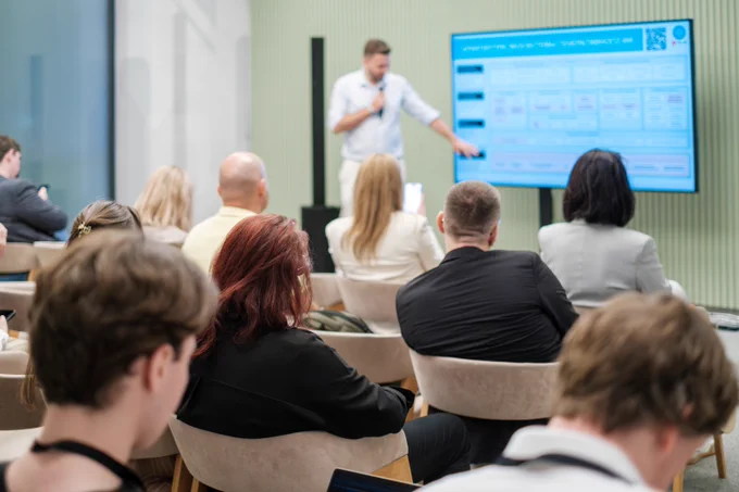 A person presenting a digital slide to an audience seated in a modern conference room.