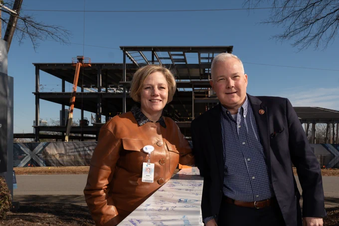 Two individuals standing in front of a construction site with signatures on a white steel beam.