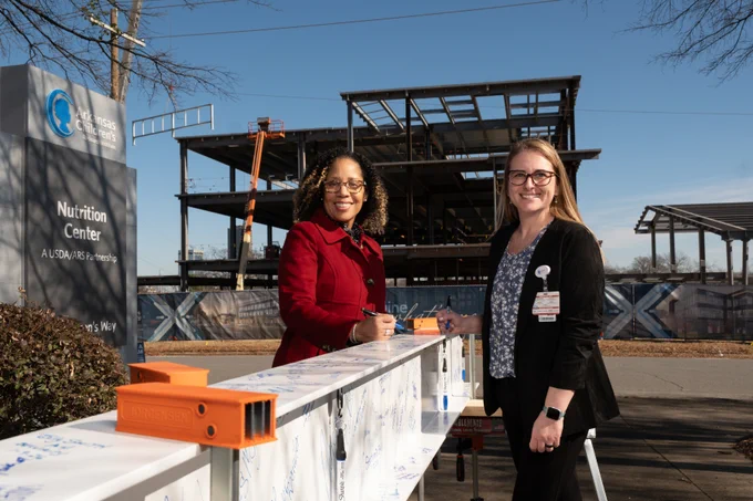 Arkansas Children's Dr. Tamara Perry and Dr. Alicia Allen standing by a beam signing table in front of the construction site for the National Center for Opioid Research & Clinical Effectiveness (NCOR).