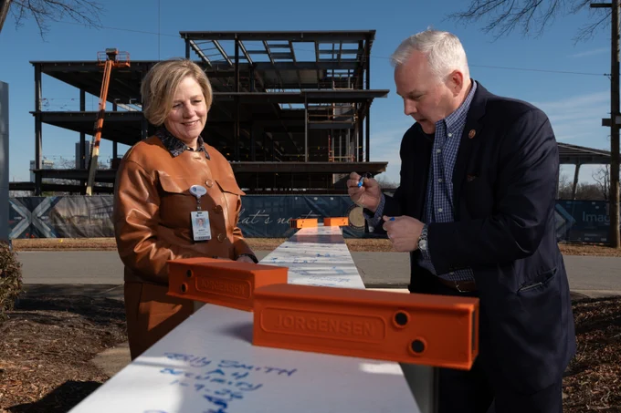 Arkansas Children's President & CEO Marcy Doderer and Arkansas Attorney General Tim Griffin signing a white beam at a construction site with a partially completed steel structure in the background. The beam features multiple signatures.