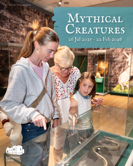 A young girl, her mother, and grandmother closely examine a coiled snake skeleton displayed in a glass case at a museum exhibit. The girl looks intrigued, pointing excitedly at the specimen, while the adults engage with her, smiling and discussing the display. In the background, a unicorn illustration and various artefacts hint at the exhibit's fantasy theme. Text in the upper right promotes the "Mythical Creatures" exhibition at Colchester Castle, running from 26 July 2025 to 22 February 2026. The Colchester Castle logo and photo credit to Tom Archer are also included.