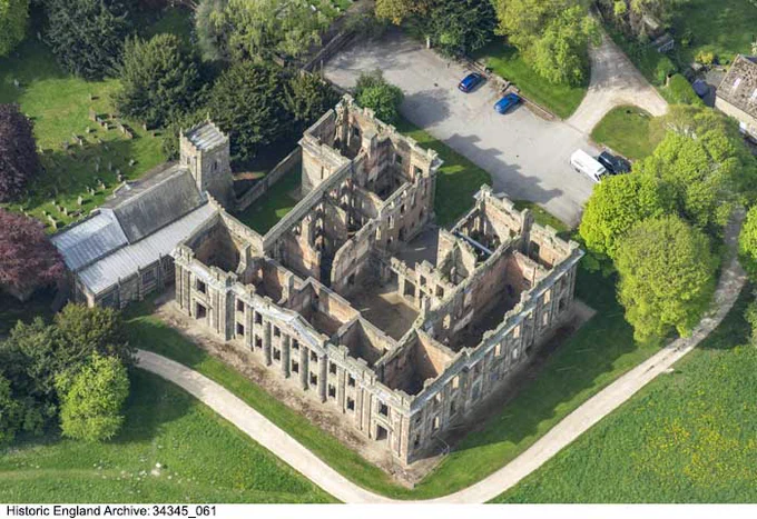 A colour photograph of an oblique aerial view of a large building without a roof, set within green trees and grass. The building is set at an angle within the photograph. At the bottom of the photograph, a pale pathway travels along two sides of the building. To the left of the building are trees and a church; to the right are also trees. To the top right of the photograph is a small car park with several cars.