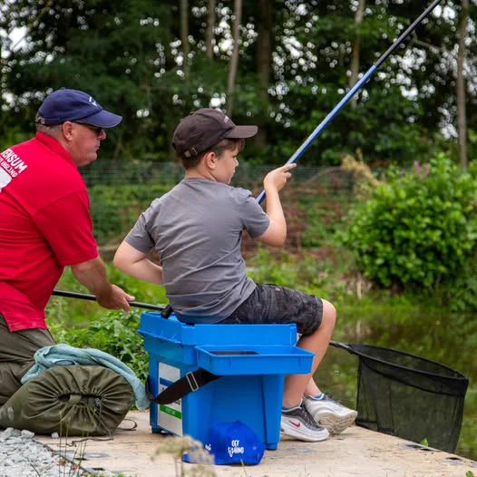 A young boy in a grey t-shirt, shorts and cap sits on a blue box on a riverbank holding a fishing rod. A man next to him in a red t-shirt and blue cap is guiding him how to fish and holding out a net under the rod and line.