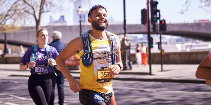 Male runner in a yellow tank top and hydration pack participating in the London Landmarks Half Marathon near a bridge on a sunny day.