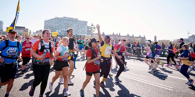 A group of marathon runners in colorful athletic gear racing on a sunny city street with spectators behind barriers.
