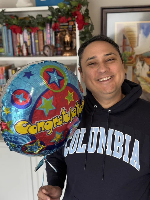 A smiling Tom wears a navy Columbia hoodie and holds a balloon that says, "Congratulations."