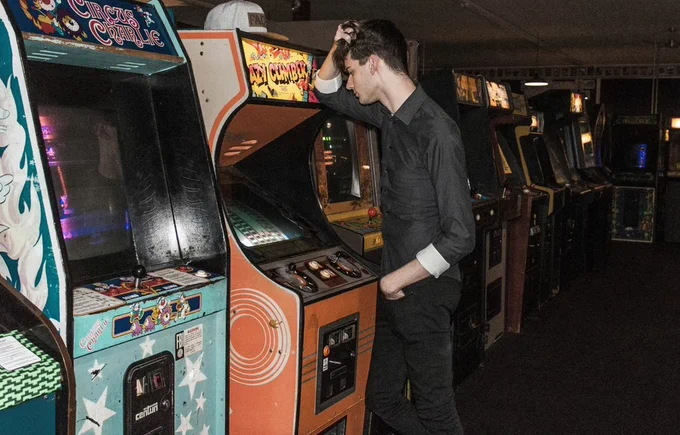 In an arcade, a guy stands leaning against the cabinet of a vintage video game. He grasps his hair in his hands, seemingly anguished by the game 'Lazy Climber'.