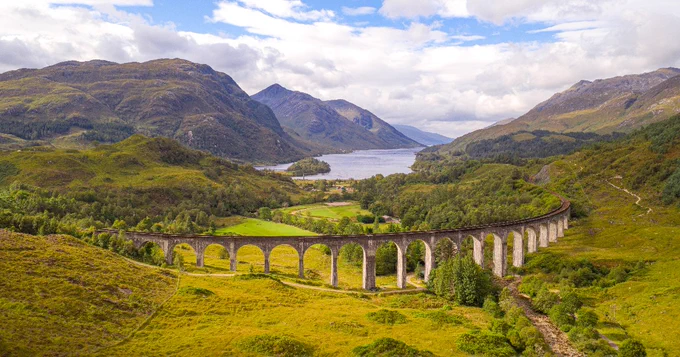 Aerial view of the Glenfinnan Viaduct in Scotland, surrounded by lush greenery and mountains with a body of water in the distance.
