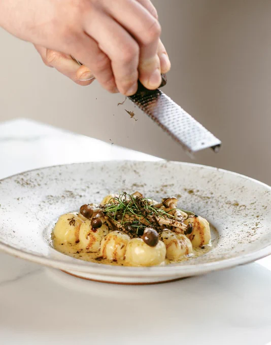 Hand grating fresh truffle over a gourmet gnocchi dish garnished with herbs and mushrooms on a white plate.