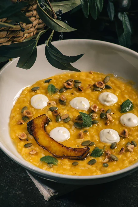 Creamy squash risotto garnished with toasted hazelnuts, pumpkin seeds, sage leaves, and dollops of white cream in a white bowl.