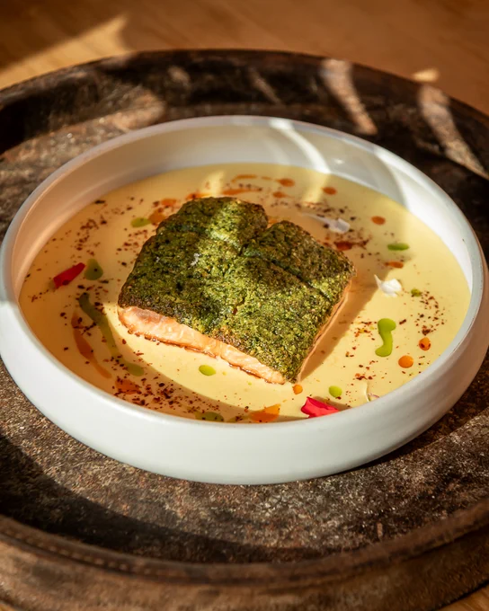 Seared salmon fillet with a green herb crust served on a creamy yellow sauce with colorful oil droplets in a white bowl.