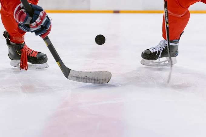 Two hockey players in red gear compete for a black puck on the ice rink during a game.