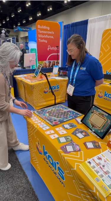 A photo of a "Kai's Education" exhibit at a trade show (FETC). A woman with long gray hair leans over a yellow table covered in educational tools, interacting with a set of directional coding cards. Standing behind the table, a company representative in a blue polo shirt and glasses looks on. The table features a colorful game board, character cards, small robots known as KaiBots, and a tablet displaying a digital interface. In the background, an orange banner reads "Building Tomorrow's Workforce, Today," alongside other convention booths.