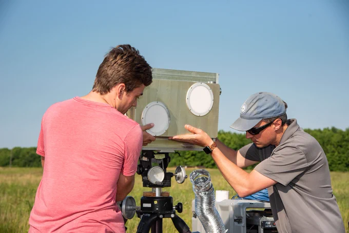 Two men adjust a metal antenna unit, approximately 12 in by 18 in, on a tripod.