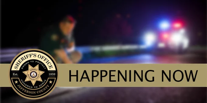 Jefferson County Sheriff’s Office alert graphic with the Sheriff’s Office logo and the words HAPPENING NOW over a blurred nighttime scene showing a deputy near police tape and flashing red and blue patrol car lights in the background.