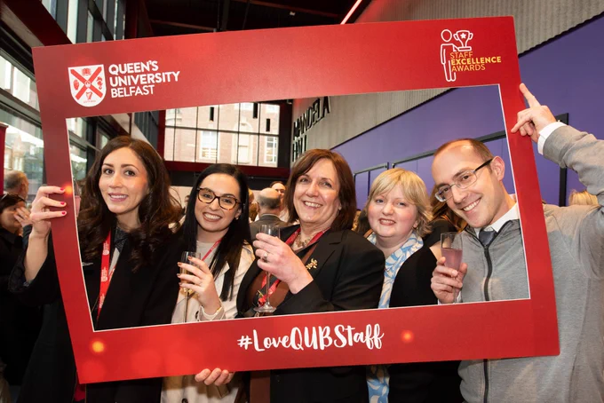 Image shows five people standing outside of the Mandela Hall in the One Elmwood Building. They are holding a red frame which has the Queen's logo on the top left-hand side, the Staff Excellence Awards logo on the top right-hand side and also has white text that reads #LoveQUBStaff along the bottom