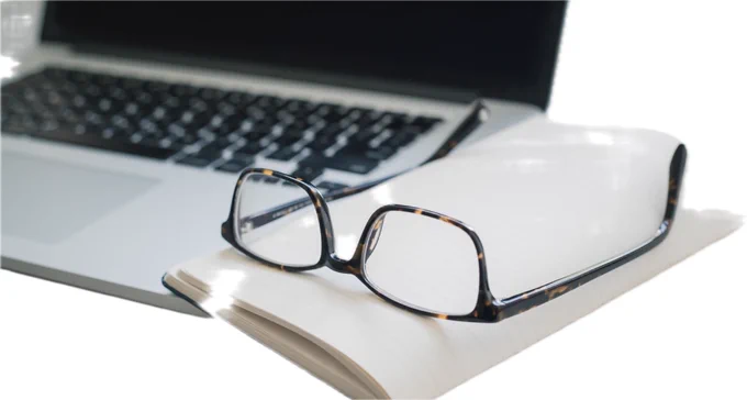 A pair of reading glasses resting on an open notebook beside a laptop, suggesting a quiet moment for reading.