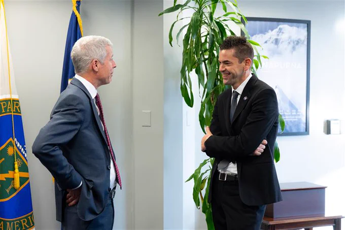 An image of Energy Secretary Chris Wright, on the left, and NASA Administrator Jared Isaacman on the right. The two men are wearing suits, facing each other, and smiling. The office-like background features two flags on stands and a tall, green plant. Credit: NASA