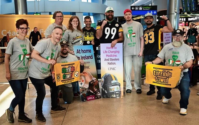 Group of people smiling, wearing Pittsburgh Steelers gear and holding signs, including one that says "Home to Life Changing Medicine. And Steelers Nation. UPMC," at an airport.