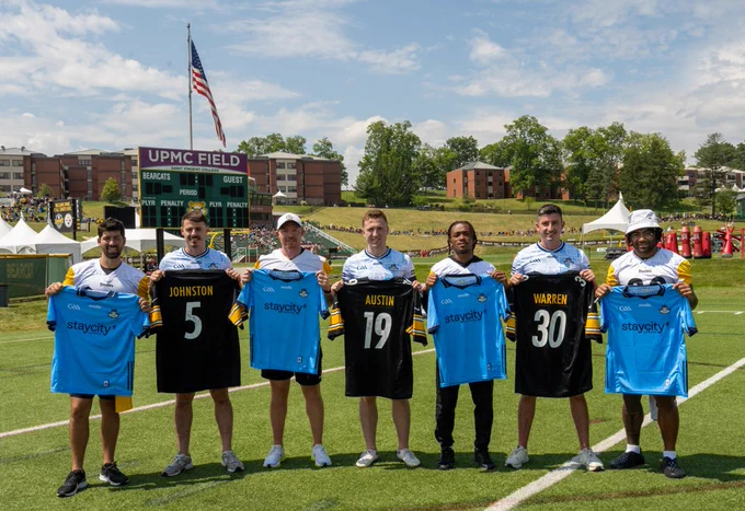 Seven athletes stand on a soccer field holding jerseys, with a scoreboard and American flag in the background. The scoreboard displays "UPMC Field."
