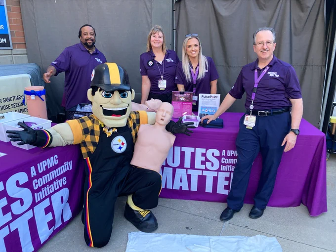 Group of individuals and the Pittsburgh Steelers mascot at a UPMC health event booth, displaying educational materials and a CPR training manikin.