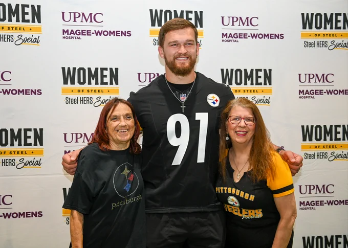 Three individuals posing together at an event with backdrops featuring logos from UPMC, Magee-Womens Hospital, and Women of Steel. The person in the middle is wearing a Pittsburgh Steelers jersey number 91.