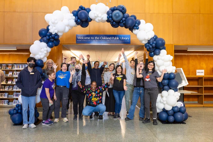 Group of people celebrating inside the Oakland Public Library, standing under a blue and white balloon arch.
