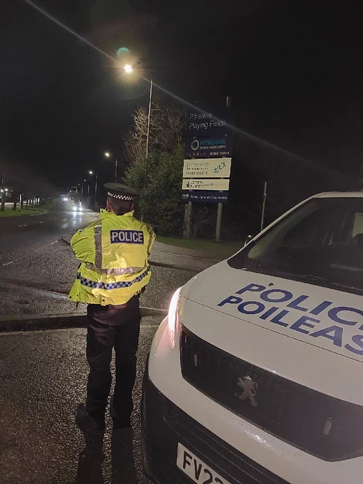 Police officer standing beside a roadway at night wearing high viz jacket holding a speed detection device. Cars with lights illuminated travelling towards the officer in the distance.