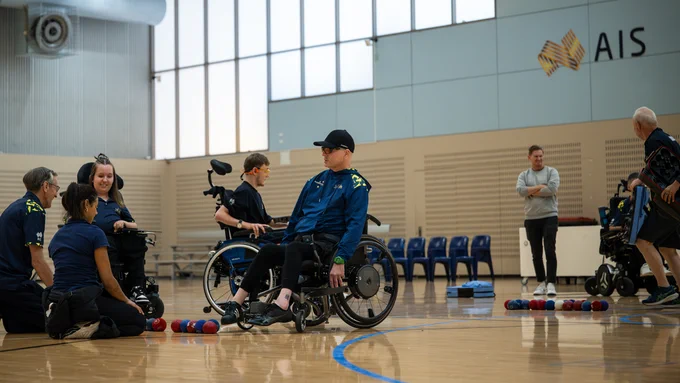 Group of Australian Institute of Sport athletes in wheelchairs and coaches discussing strategy on a gym floor with boccia balls nearby.