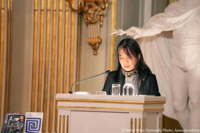 Han Kang delivering her Nobel Prize lecture at the Swedish Academy, in a room with ornate golden columns and curtains.