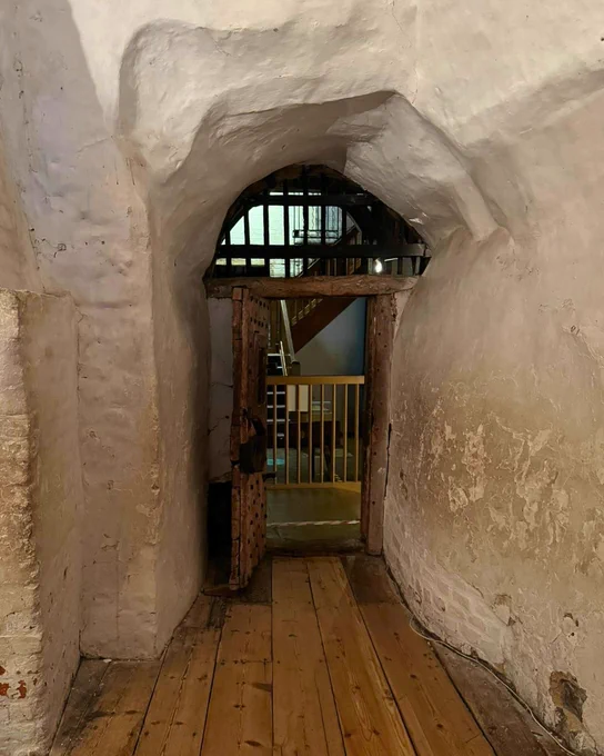 A dim stone corridor in the castle prisons, with heavy brick walls, a low arched ceiling and metal-barred doors on either side. The passage is empty and lit by a few warm wall lights, so the far end of the corridor fades into shadow, giving the scene a tense, watchful atmosphere.
