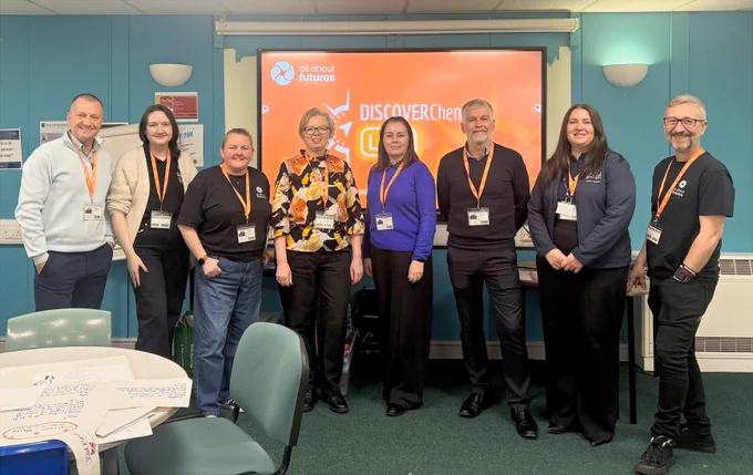 Members of All About Futures with delivery partners from CATCH, Education Academy Scotland, and RTC North. The teams are preparing to host IChemE workshops in a classroom and there is a blue wall with a digital whiteboard behind them. The whiteboard shows an orange background with the All About Futures and IChemE DiscoverChemEngLIVE logos on it in white.