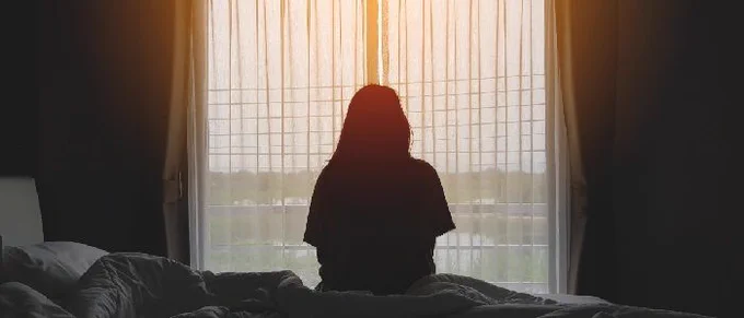 Silhouette of a person sitting in bed with curtains and a sun peering through the window 