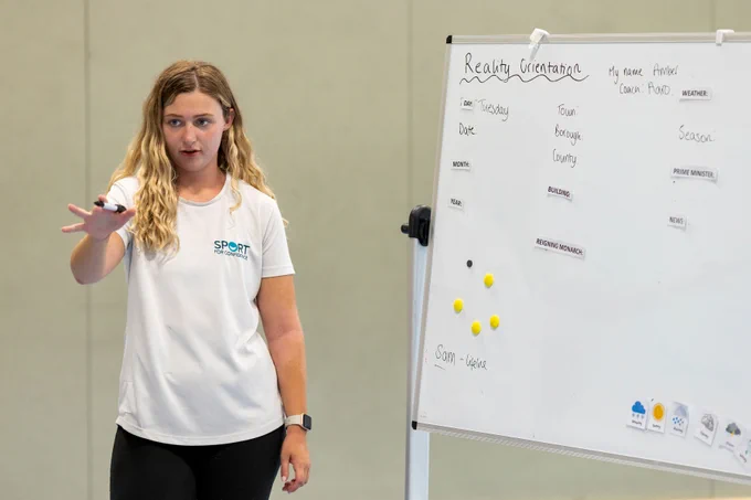 A female Sport for Confidence staff member standing by a whiteboard, a black whiteboard marker raised in her hand as she talks to a group of people who are out of shot.