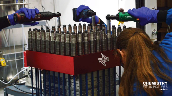 A woman in a lab coat uses a rotary tool to tighten one of a series of about 35 rods going around the perimeter of a stack of metal plates that make up an electrochemical stack. The hands and tools of several other people can be seen tightening other rods.