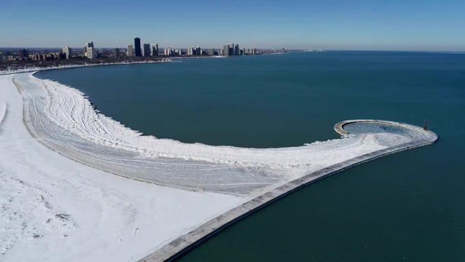 Aerial view of Montrose Beach jetty in winter ice and snow accumulating over the sandy shore and northern Chicago neighborhoods in the distance.