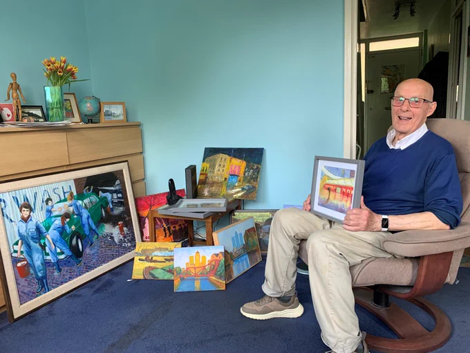 white man in his 60s sitting with painting on his lap, in a room with dark blue carpets and light ble walls, with smaller paintings on the floor and a larger one against a couple of chest of drawers