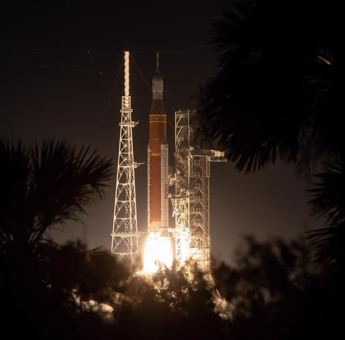 NASA’s SLS (Space Launch System) rocket carrying the Orion spacecraft launches on the Artemis I flight test on Nov. 16, 2022, from Launch Complex 39B at NASA’s Kennedy Space Center in Florida. The rocket has an orange core stage and two white boosters on either side. The glow underneath the rocket is illuminating the night sky. Palm trees in silhouette in the foreground partially cover the view. Credit: NASA/Bill Ingalls