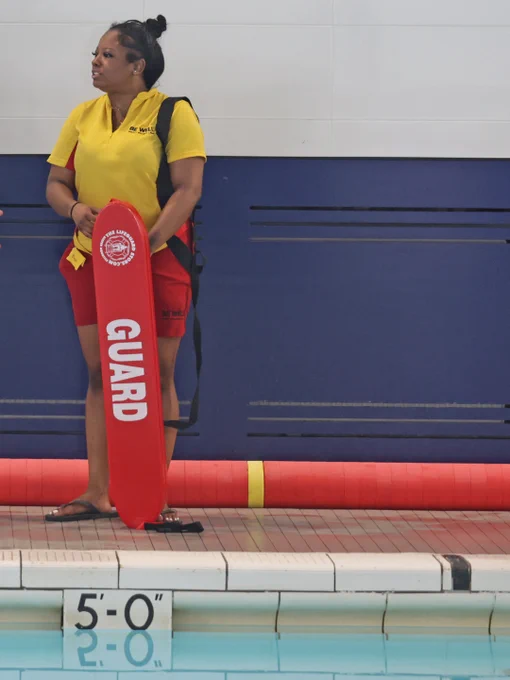 A photo of a female black lifeguard standing by the edge of a pool. She is in red and yellow lifeguard uniform.