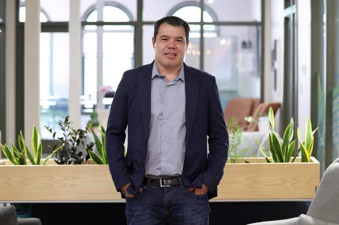 A man with short black hair wears a suit and shirt with jeans and his hands in his pockets. There is a blurred background behind him of windows and plants.