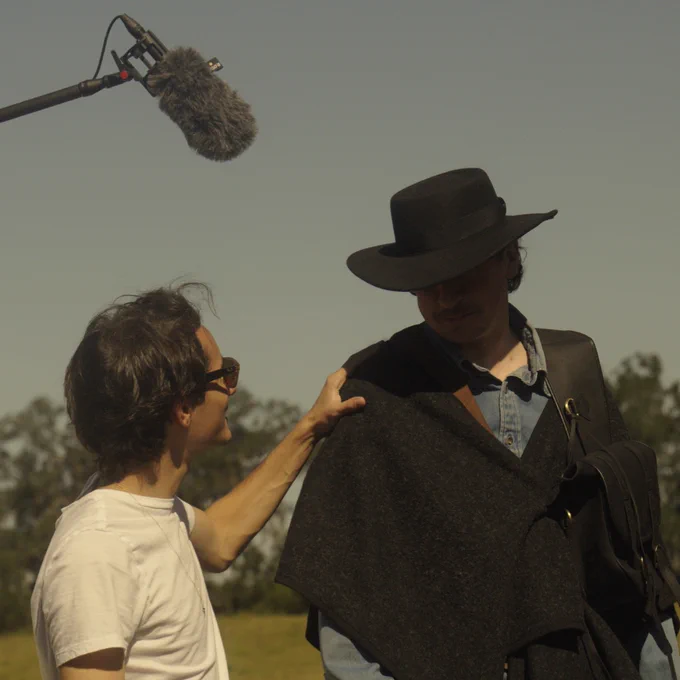 Person speaks with another individual wearing a hat and historical outfit, with a microphone boom overhead, outdoors.
