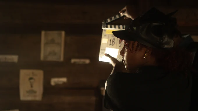 A person operates a clapperboard on a dimly lit film set, with the backdrop featuring rustic wooden walls and vintage posters.