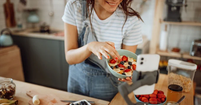 Young woman in apron preparing a colorful fruit bowl while recording a cooking video on her smartphone in a cozy kitchen.